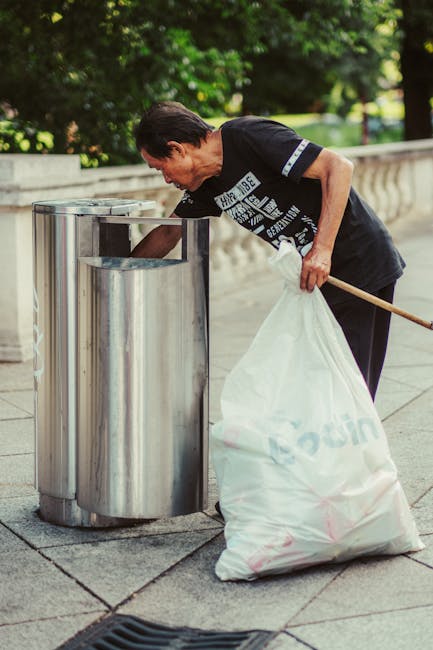A middle-aged man with dark hair, dressed in a black T-shirt and dark trousers, is bent over a silver metal public rubbish bin on a paved sidewalk. He is holding a long-handled litter picker in his right hand and has a large white plastic bag, which appears to be partially filled with collected debris, in his left hand. The metal bin has a cylindrical shape with a slot for inserting waste and is situated outdoors, with a background of lush green trees and a stone railing, suggesting a park or landscaped urban area. The scene is illuminated by natural daylight, casting soft shadows on the ground. The image depicts active waste collection in an outdoor environment, reflecting services like private rubbish removal or on-site clearance, with the man engaged in clearing litter from the public space, consistent with the practices of a waste disposal company like Waste Disposal Ealing.