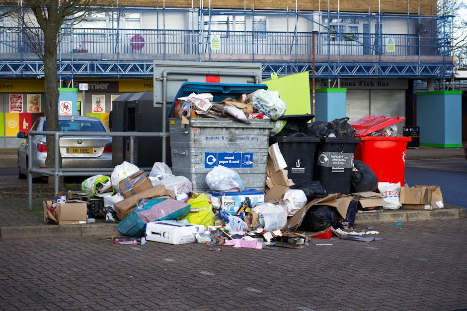 A collection of overflowing rubbish bins and scattered waste on a paved sidewalk area, with a large grey bin filled with mixed paper and cardboard stacked on top of it, surrounded by black and red wheelie bins for general waste disposal. Cardboard boxes, plastic bags, and loose paper are strewn across the ground, indicating overfilled containers. In the background, a street scene features parked cars, including a silver vehicle, and a commercial building with storefronts and signs, some covered in blue scaffolding. The scene appears to be an outdoor urban environment during daylight, with the waste accumulation suggesting a need for rubbish removal services. The setting highlights the importance of private refuse collection or on-site clearance for maintaining cleanliness in public spaces, as managed by waste disposal companies such as Waste Disposal Ealing.