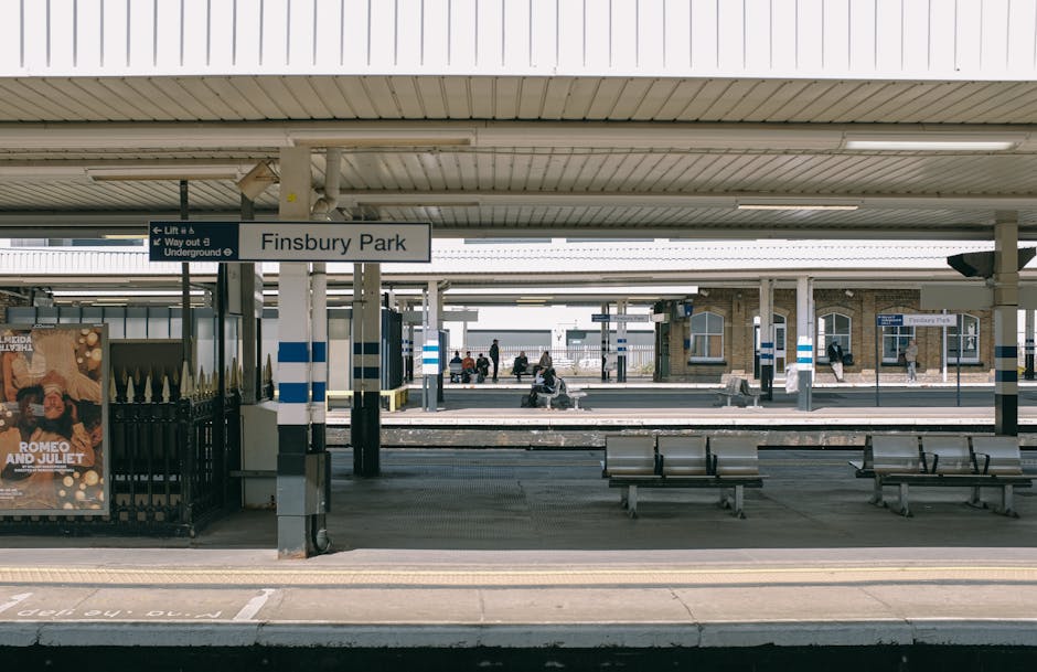 View of Finsbury Park station platform showing multiple wooden benches with metal frames positioned along the edge of the platform in the foreground. The platform is covered by a large, corrugated metal roof supported by evenly spaced metal columns, some featuring blue and white striped markings. In the middle ground, several passengers are seated on the benches or standing, waiting for a train, with some looking at their phones or talking. To the left, a black metal fence encloses a small advertisement display for a theatrical production of Romeo and Juliet, with a colorful poster visible. The background shows additional platforms, tracks, and an older brick station building with arched windows, as well as a few more waiting passengers. The scene is lit by natural daylight, creating a neutral and calm atmosphere, reflecting a typical urban railway station setting, relevant to discussions of public transport and alternative arrangements for mobility or travel in the London area.