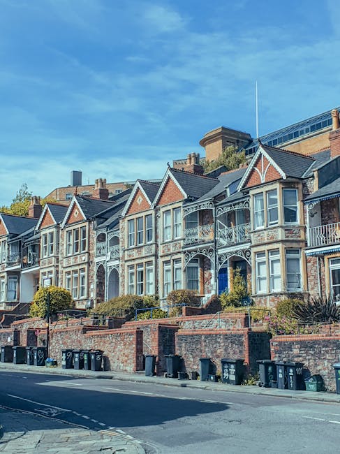 A row of Victorian-style terraced houses with ornate Victorian ironwork balconies and decorative gables, situated on a slightly elevated street level with a brick retaining wall at the front. The houses have light-colored window frames and brick facades with contrasting detailing, set against a clear blue sky. In the foreground, there are several black wheelie bins lined up along the pavement, indicating waste collection or disposal services. The scene appears to be in a residential area that may require alternative waste handling, with Waste Disposal Ealing potentially providing independent rubbish removal or on-site clearance for the properties. The environment suggests a suburban setting typical of London neighborhoods, emphasizing private rubbish management over municipal collection. The lighting is natural and bright, highlighting the architectural details and the tidy appearance of the street, with lush greenery visible above the rooflines and along the staircase pathways leading to each front door.