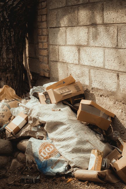 A pile of mixed waste materials situated at the base of an exterior brick wall, partially shaded by a tree to the left. The waste includes flattened cardboard boxes of various sizes, some with printed labels and barcode stickers, along with plastic and paper packaging. A large textured white sack is visible among the rubbish, partially covering some of the cardboard. The ground consists of dirt and small rocks, with some debris scattered around. The brick wall behind displays light-colored, weathered bricks, and the scene suggests an informal or partial rubbish accumulation likely intended for collection or disposal. The scene may relate to private waste handling or alternative rubbish removal services, such as those provided by Waste Disposal Ealing, for on-site clearing or non-contracted collection in an urban or suburban setting.