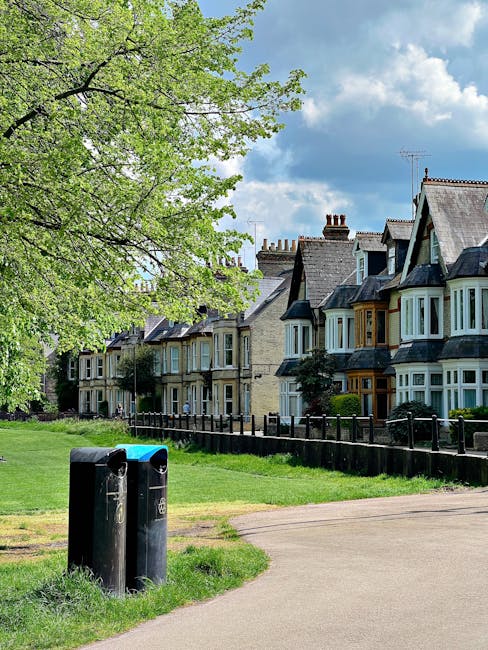 A row of Victorian-style terraced houses with ornate Victorian ironwork balconies and decorative gables, situated on a slightly elevated street level with a brick retaining wall at the front. The houses have light-colored window frames and brick facades with contrasting detailing, set against a clear blue sky. In the foreground, there are several black wheelie bins lined up along the pavement, indicating waste collection or disposal services. The scene appears to be in a residential area that may require alternative waste handling, with Waste Disposal Ealing potentially providing independent rubbish removal or on-site clearance for the properties. The environment suggests a suburban setting typical of London neighborhoods, emphasizing private rubbish management over municipal collection. The lighting is natural and bright, highlighting the architectural details and the tidy appearance of the street, with lush greenery visible above the rooflines and along the staircase pathways leading to each front door.