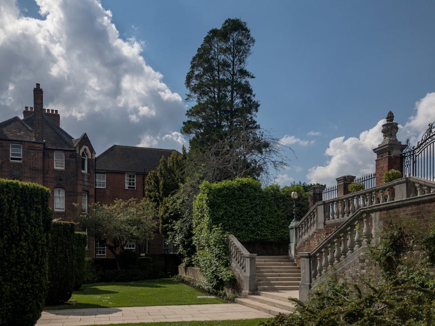A set of outdoor stone stairs with a decorative balustrade and ornate pillars leading up to a gated entrance on a residential property. Surrounding the stairs are lush green bushes and trees, with a tall, slender chimney visible on a nearby brick building. The area appears to be part of a well-maintained garden or landscaped yard, bordered by a paved pathway at the base of the stairs. The sky above is partly cloudy with patches of blue, indicating a bright day, ideal for outdoor maintenance or private waste clearance. The scene is quiet and orderly, reflecting typical garden scenery in an upscale residential area, without visible rubbish or debris, and subtly aligns with the context of private waste management or garden clearance services provided by Waste Disposal Ealing.