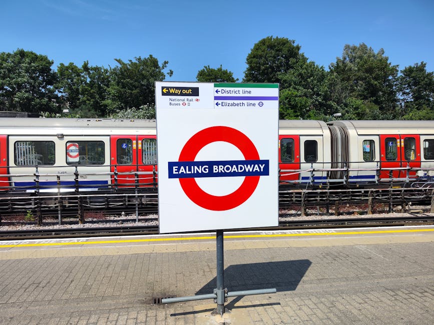 A white sign with a red circular symbol and a blue horizontal band displaying 'EALING BROADWAY', positioned on a paved station platform. The sign features directional information with black and purple text, indicating routes and lines including the National Rail, District line, and Elizabeth line, along with an arrow pointing left labeled 'Way out'. Behind the sign, a modern train with a silver body and red, white, and blue exterior is visible, parked along the railway tracks. The train's exterior shows glass windows and doors, with some reflections and interior glimpses. To the left of the train, metal fencing runs parallel to the tracks, providing safety barriers. In the background, a dense row of leafy green trees and bushes extends across the top of the image, under a clear blue sky. The lighting suggests a bright, sunny day, casting defined shadows on the platform. This scene, captured at Ealing Broadway station, reflects the urban transport infrastructure that facilitates efficient passenger movement and the multimodal transit options serving the area, aligning with the concept of alternative transportation and potentially private or independent collection methods provided by waste management services in the area.