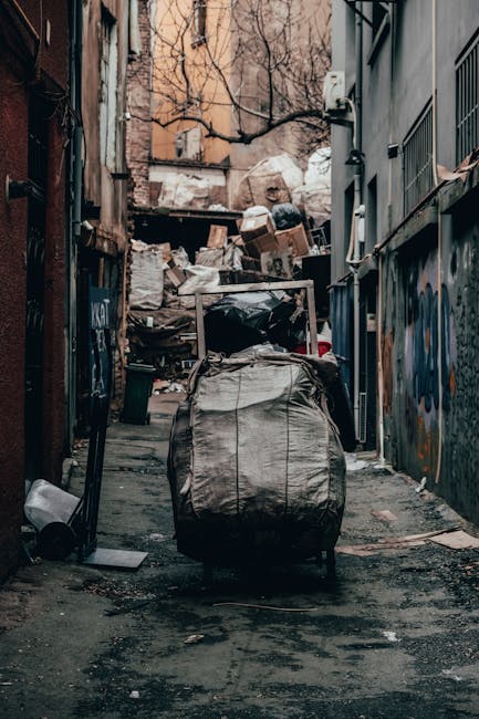 A narrow alleyway filled with a large, wheeled waste collection trolley positioned in the foreground, covered in a weathered, grey fabric material. In the background, a pile of mixed waste, including cardboard boxes, plastic bags, and other refuse, is stacked against a wall of a building with peeling paint and graffiti. To the right, a dark grey building with metal bars on the windows and a small air conditioning unit mounted near the top is visible. The ground is uneven, stained with dirt and grime, and scattered with debris, indicating an area used for private waste disposal rather than official refuse collection. The scene appears to be in an urban environment, with bare tree branches overhead and additional multi-storey buildings in the distance, suggesting an alley used for rubbish removal or on-site clearance, consistent with the services offered by Waste Disposal Ealing.