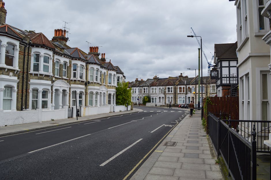 A quiet residential street lined with traditional Victorian-style terraced houses featuring brick façades, white decorative trims, and large bay windows. The buildings are mostly three to four storeys tall, with tiled, sloped roofs and prominent chimney stacks. On the right side of the image, there is a sidewalk paved with concrete slabs, accompanied by a low black metal railing in front of cream-colored, multi-story homes. The street is void of parked vehicles on the left side, with a few cars visible further down. A solitary pedestrian is walking along the sidewalk towards the background, where additional rows of similar houses can be seen. Tall street lamps with modern fixtures are positioned intermittently along the pavement, and an under-construction crane is visible in the distance against a cloudy grey sky, suggesting ongoing development in the area. The scene appears to be during daytime with diffused natural light, typical of overcast weather. This urban environment exemplifies typical London residential architecture, where private waste disposal services like Waste Disposal Ealing often manage rubbish clearance for such properties.