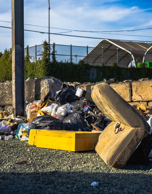 A collection of mixed waste including black rubbish bags, discarded packaging, and a worn out car seat is piled on a gravel surface outdoors in front of a low stone wall. The waste is cluttered and appears to be a small-scale rubbish clearance, possibly from a household or garden. Behind the wall, there are green bushes and a building with a curved, semi-open metal roof structure. Overhead, a utility pole with attached power lines spans the scene, which is lit by natural daylight, creating clear shadows on the gravel. The overall setting suggests an informal rubbish collection point, potentially managed by Waste Disposal Ealing for alternative waste handling or private disposal services, rather than standard bin collection. The scene emphasizes the need for professional rubbish removal services to manage such clutter effectively.